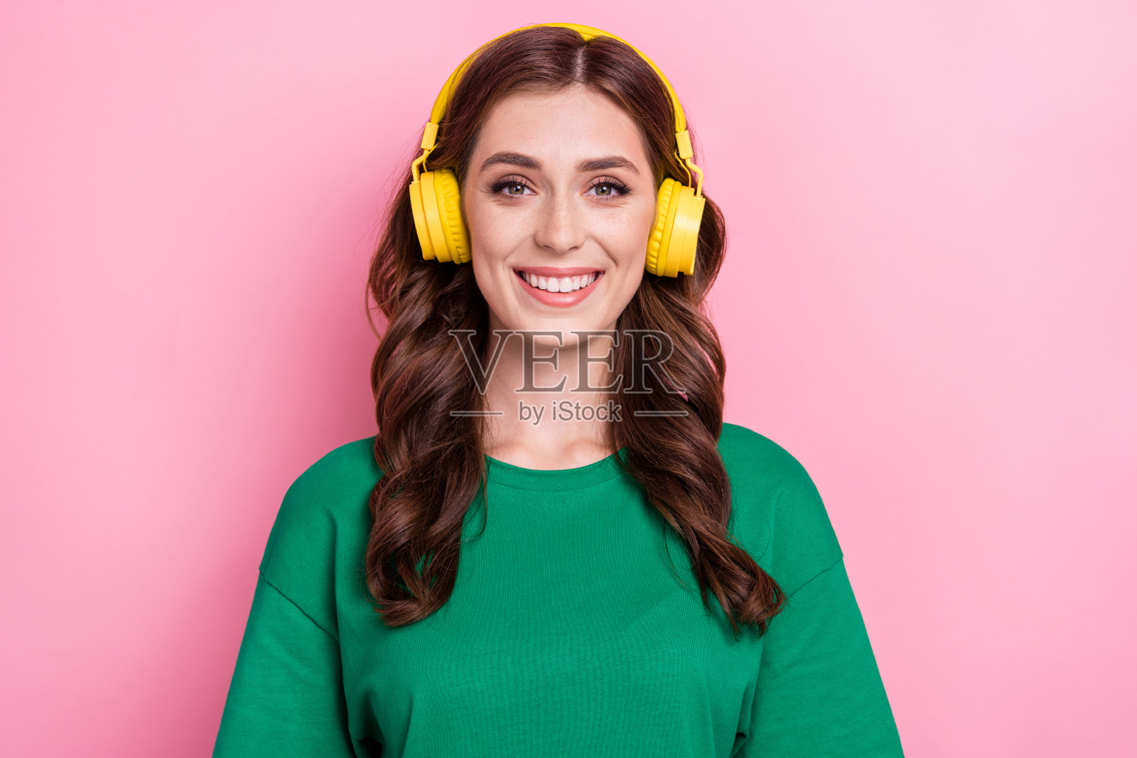 一位愉快的女性,留着波浪卷发,穿着绿色T恤,戴着耳机,正在聆听她最喜欢的音乐,背景为粉色。照片摄影图片