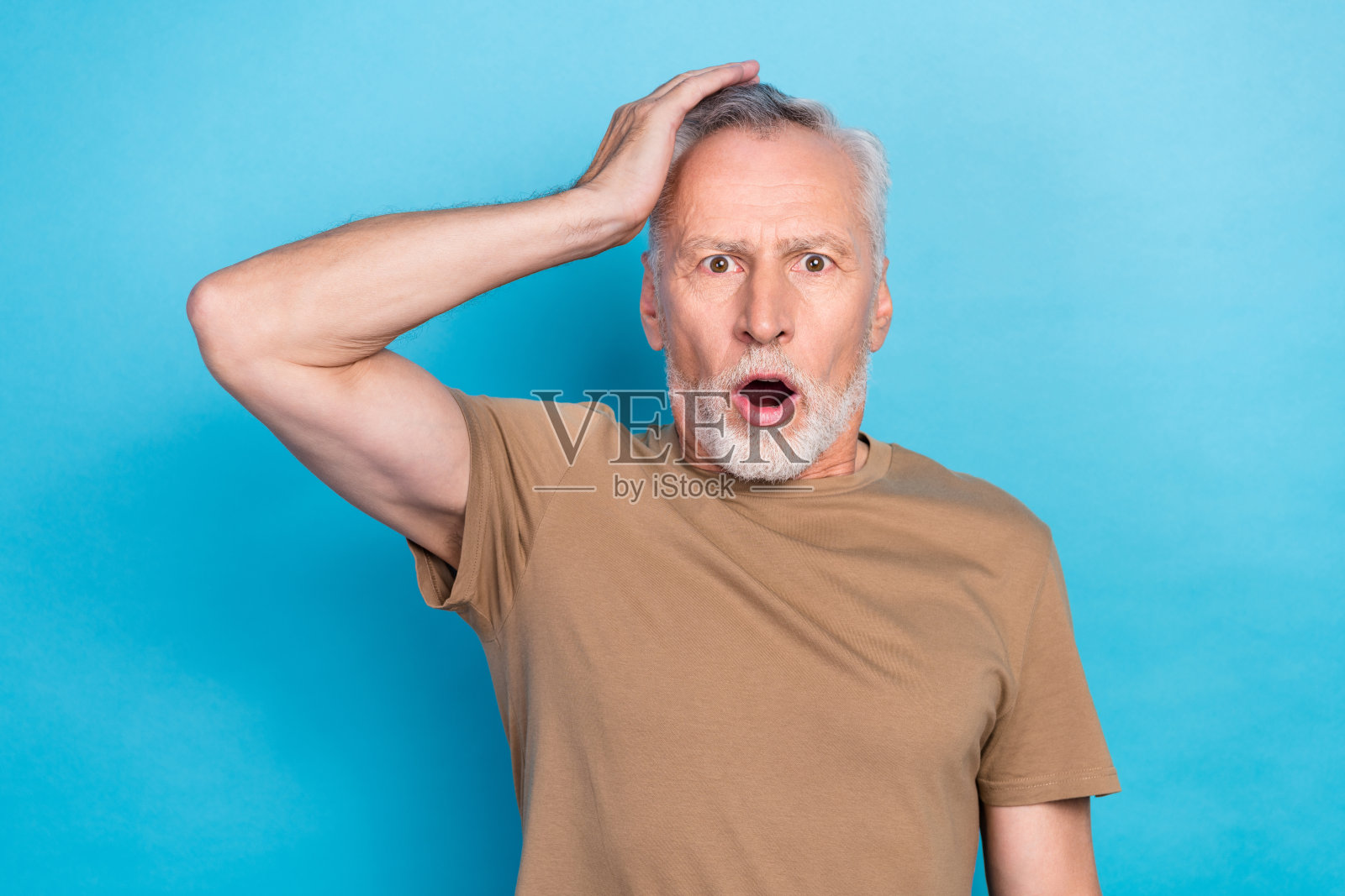 一位无言且印象深刻的老年男子,灰色发型,胡茬,穿着米色T恤,手掌放在头上,背景为蓝色。照片摄影图片