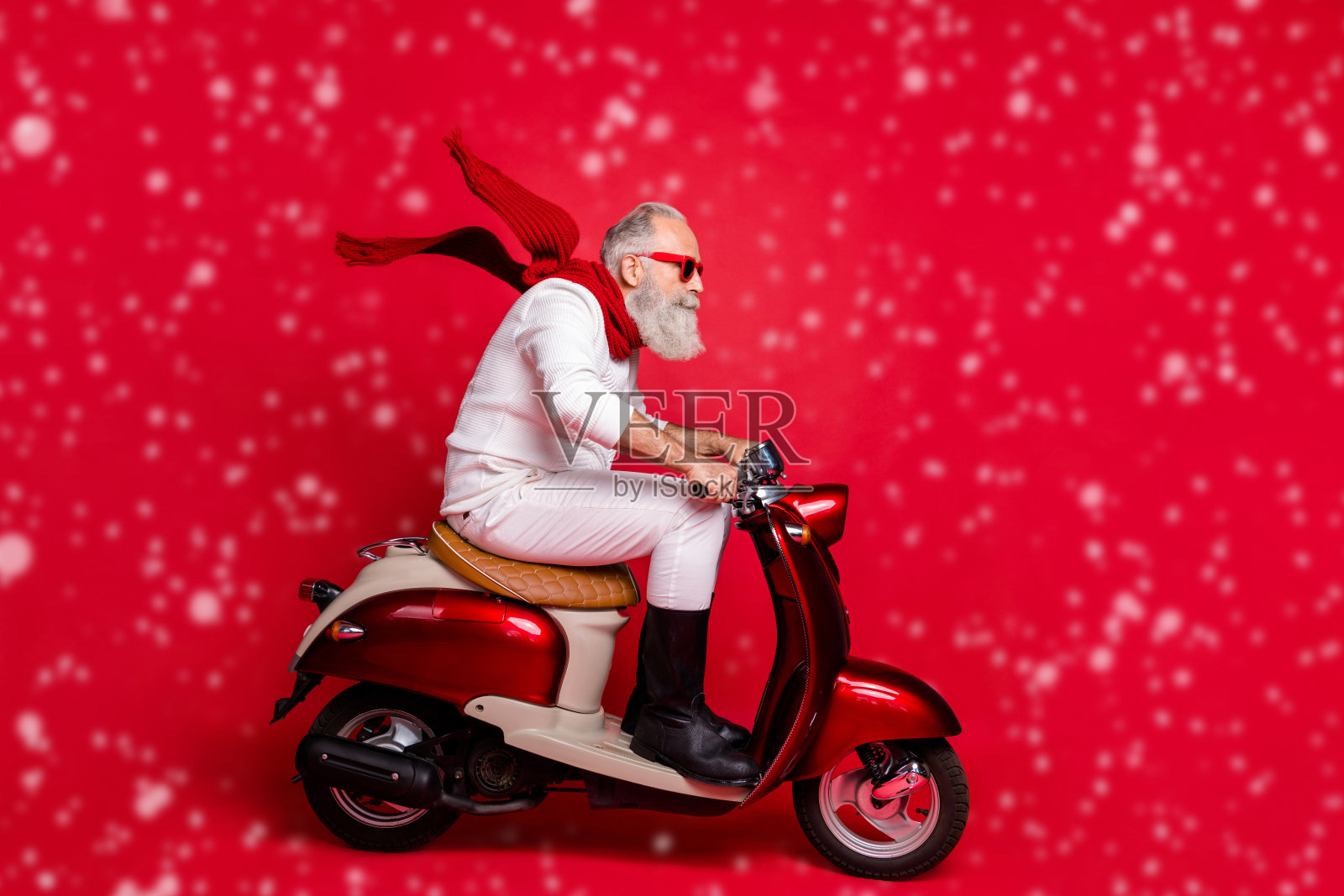 酷炫的老圣诞老人骑着自行车赶往新年派对,戴着太阳镜,穿着毛衣和裤子,背景为红色。照片摄影图片