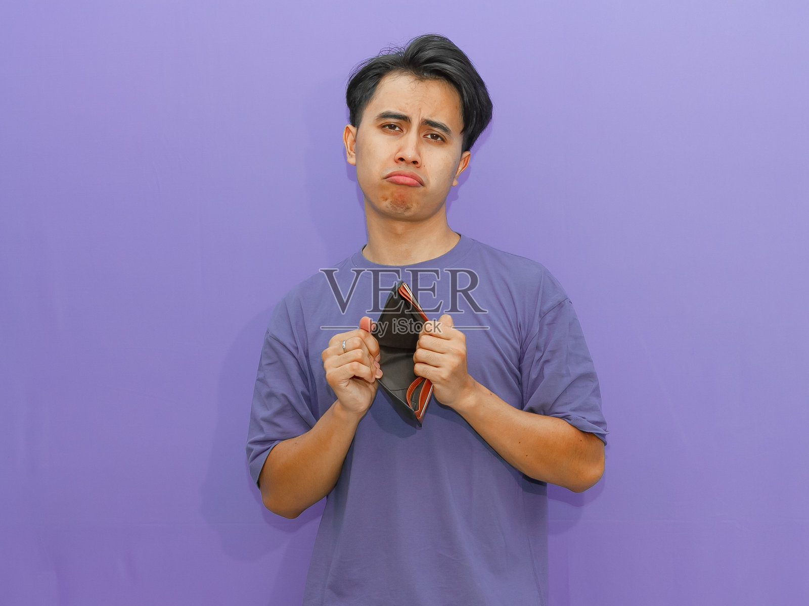 忧伤的年轻亚洲男子手里拿着空钱包,面露压力,背景为紫色,表现出经济困境和艰辛。照片摄影图片