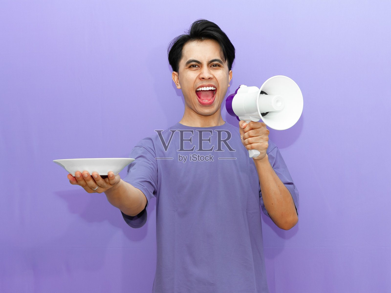 兴奋的年轻亚洲男子手持空盘,通过扩音器大喊,背景为鲜艳的紫色,非常适合食品广告。照片摄影图片