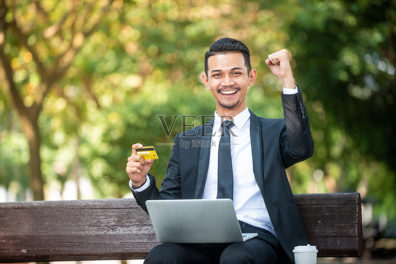 年轻快乐的商人坐在长椅上,手持信用卡,使用笔记本电脑,背景是树木。照片摄影图片