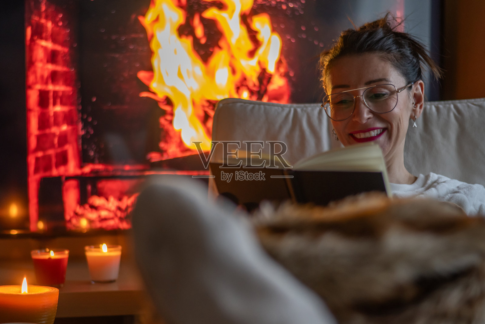女性,快乐而微笑,享受在温馨冬季环境中靠近壁炉阅读书籍的放松时光。完美的闲暇时光,放松身心,远离喧嚣。照片摄影图片