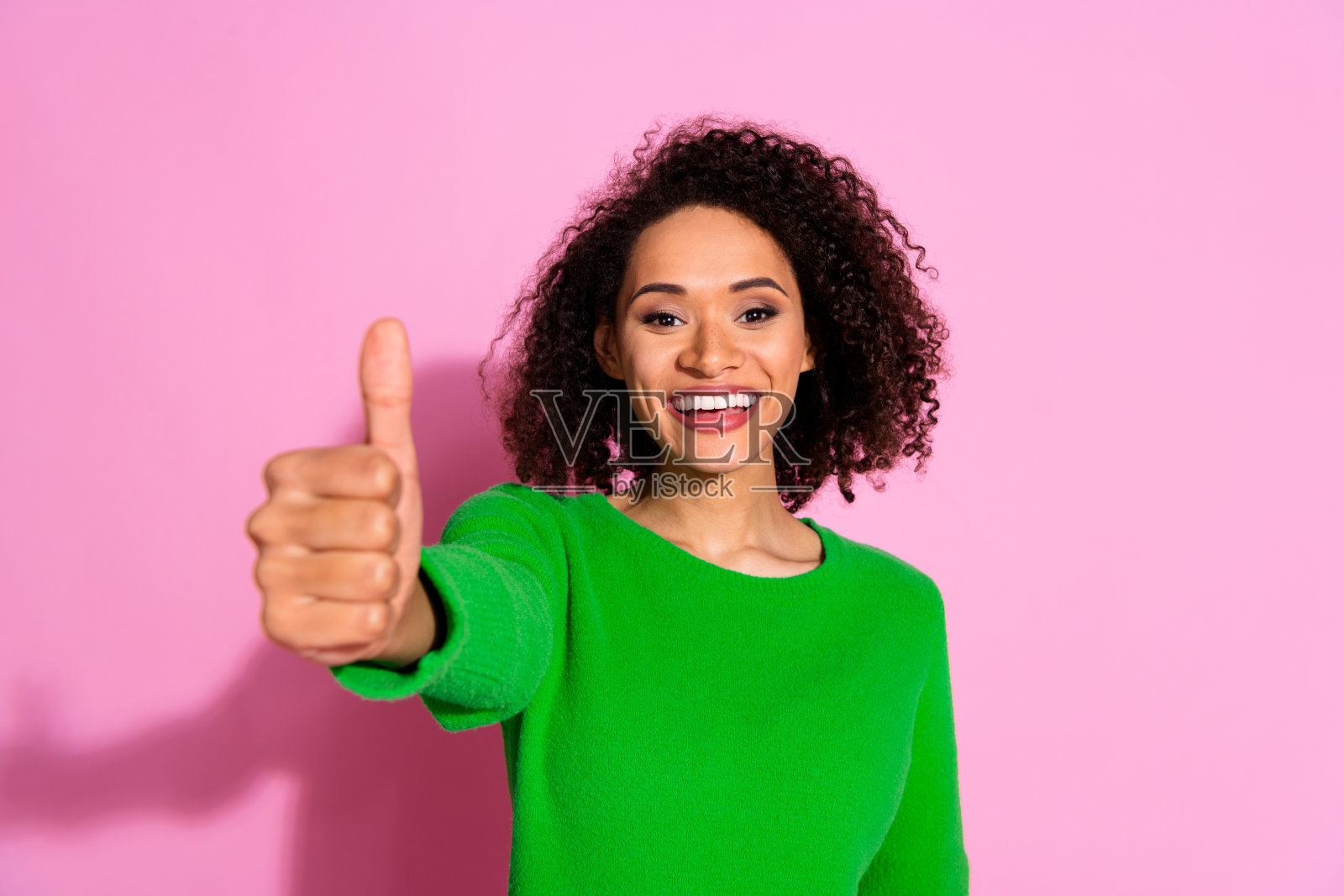 穿着绿色时尚服装、面带微笑的可爱女性在粉色背景上竖起大拇指。照片摄影图片