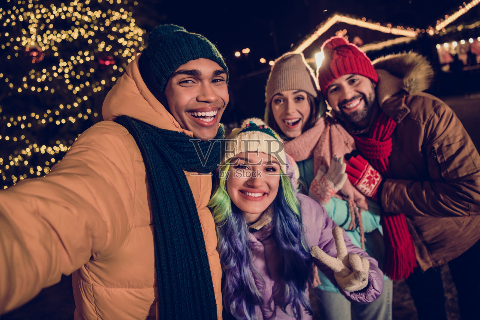 四个酷炫的朋友穿着风衣,在城市市场公园外一起录制圣诞视频博客,做出V字手势。照片摄影图片