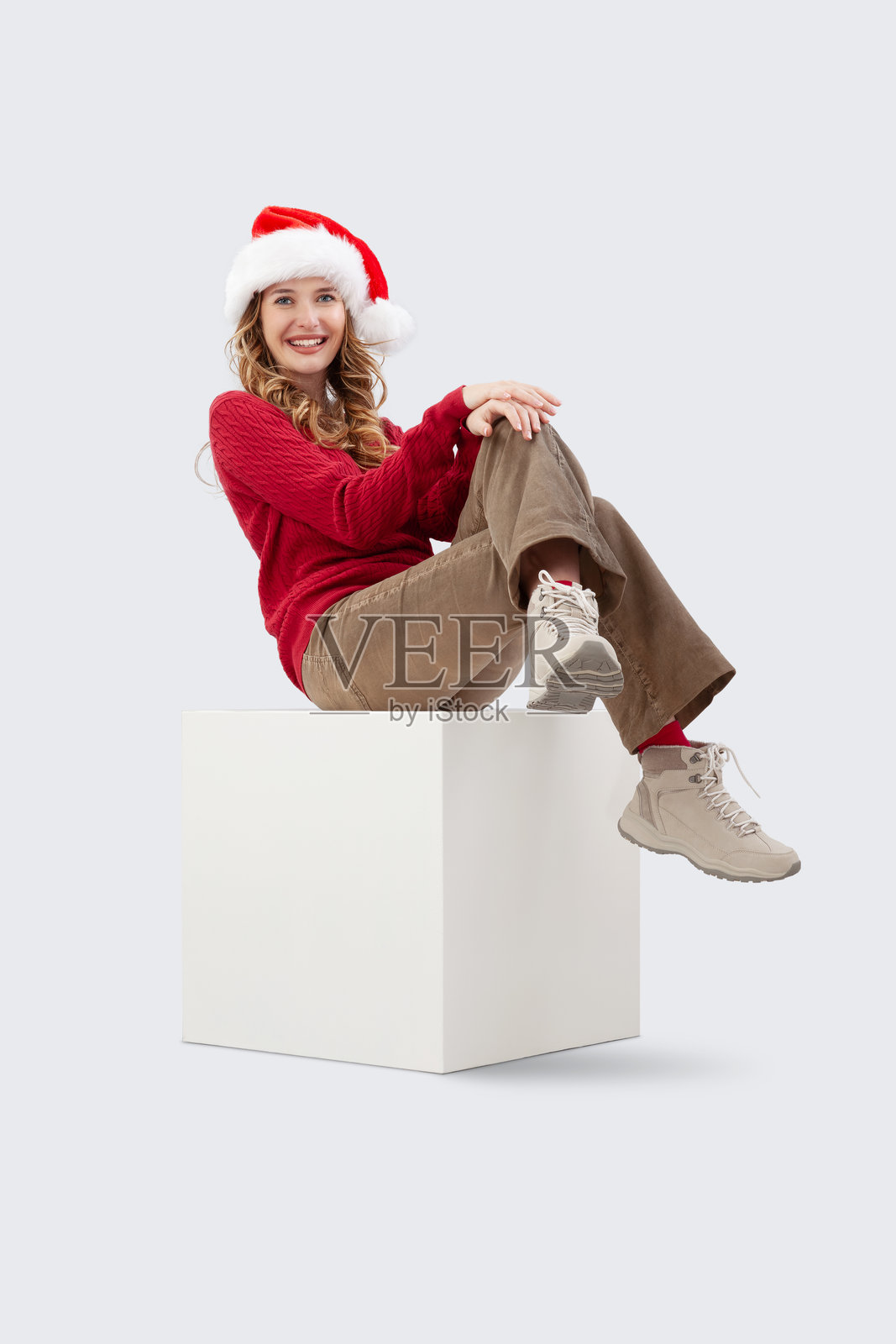 圣诞快乐。一位美丽微笑的年轻金发女性坐在一个白色立方体上,面向镜头。她戴着圣诞老人帽,穿着红色毛衣,背景为白色。照片摄影图片