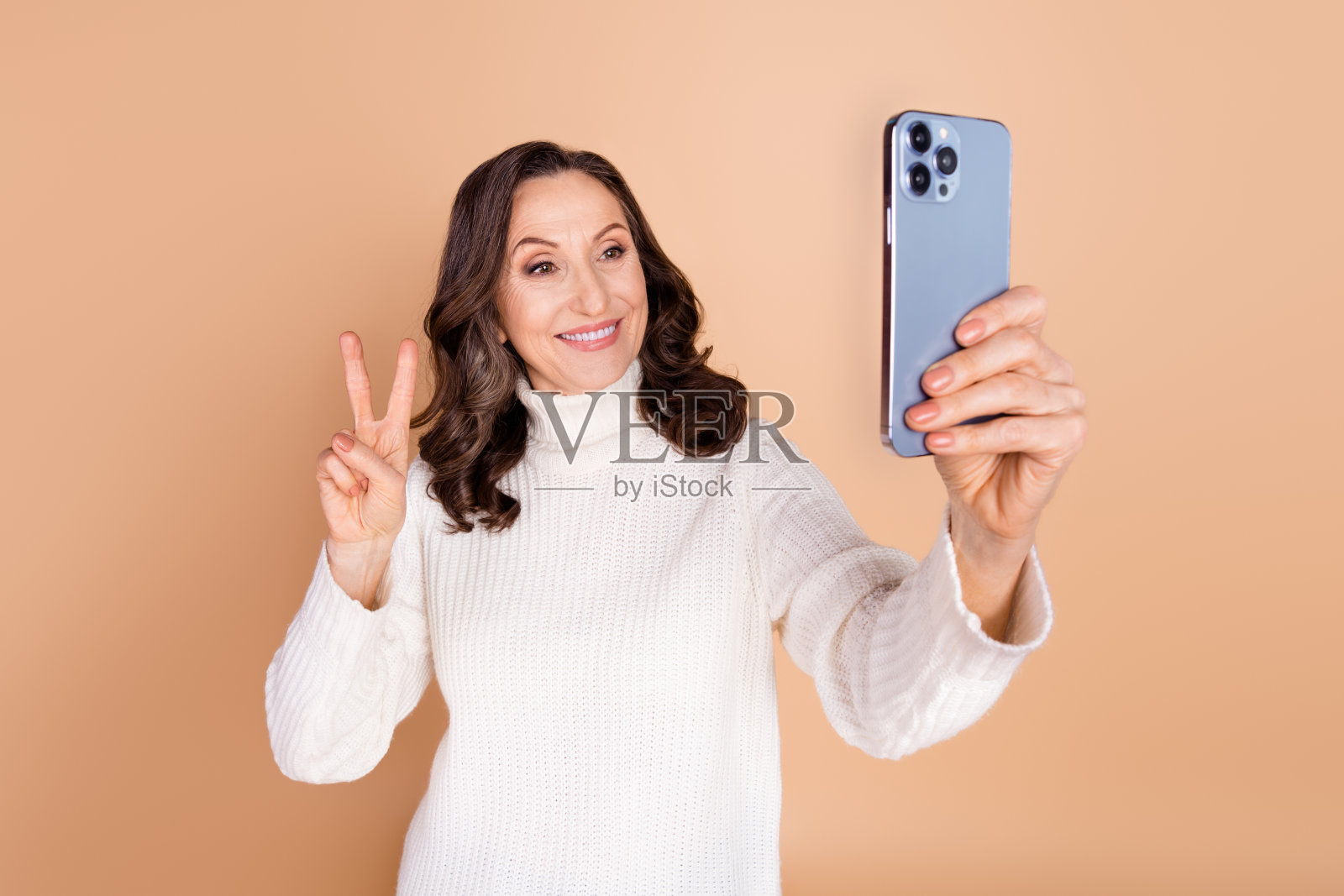 穿着白色时尚服装的迷人可爱的老年女性,手持电子设备,做出V字手势,背景为米色。照片摄影图片