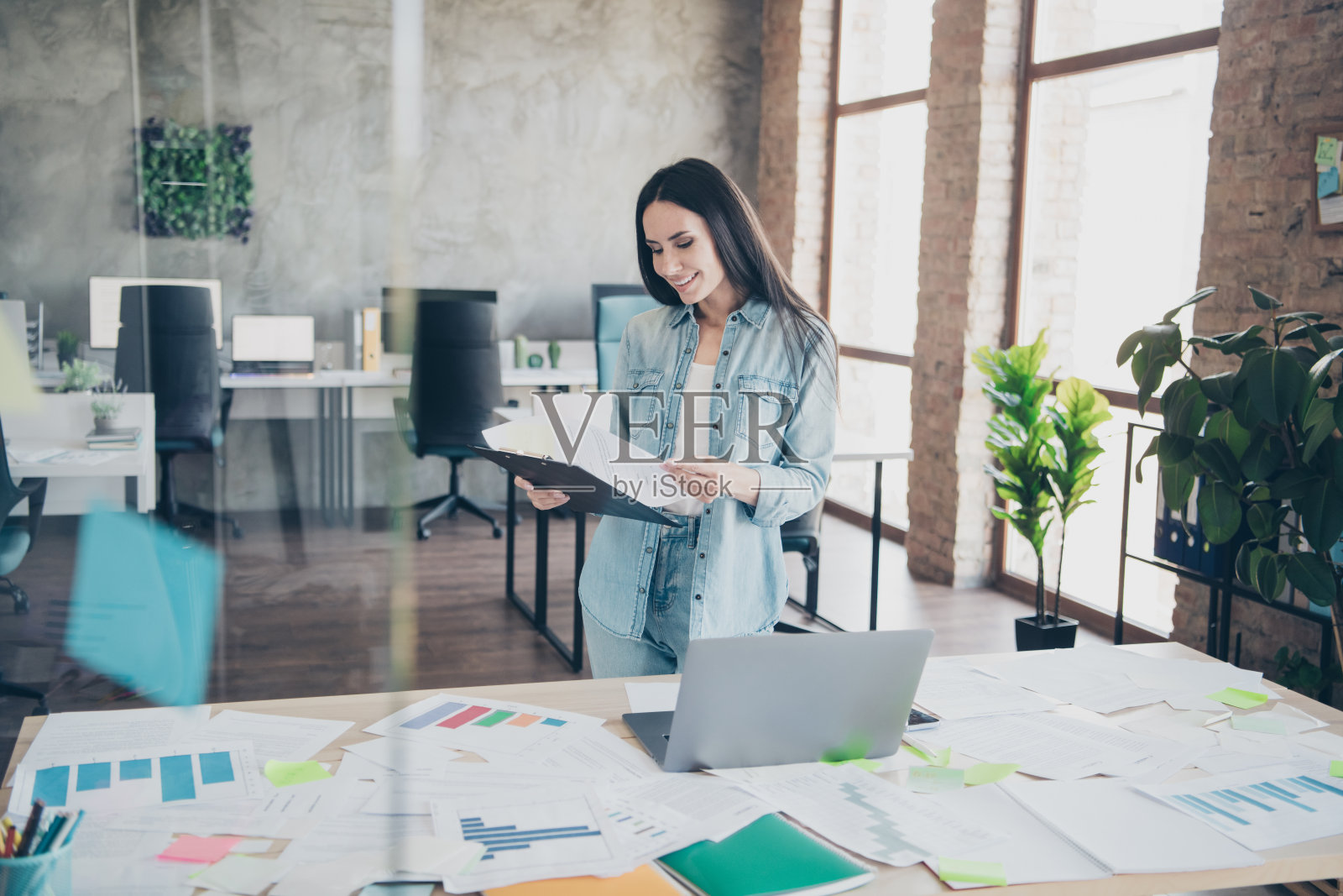 时尚牛仔服装的可爱年轻女士在现代办公环境中阅读文件照片摄影图片