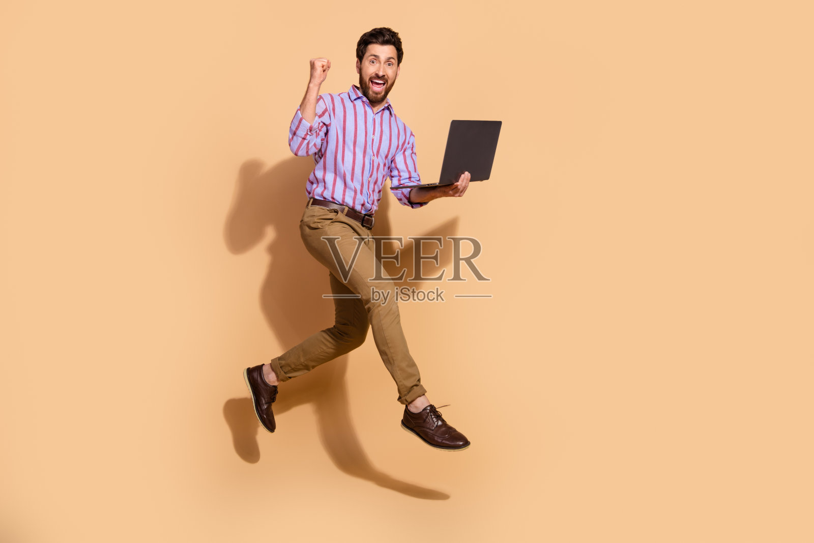 棕发成年男性跳跃,办公室经理举起拳头庆祝虚拟项目的胜利,背景为米色。照片摄影图片