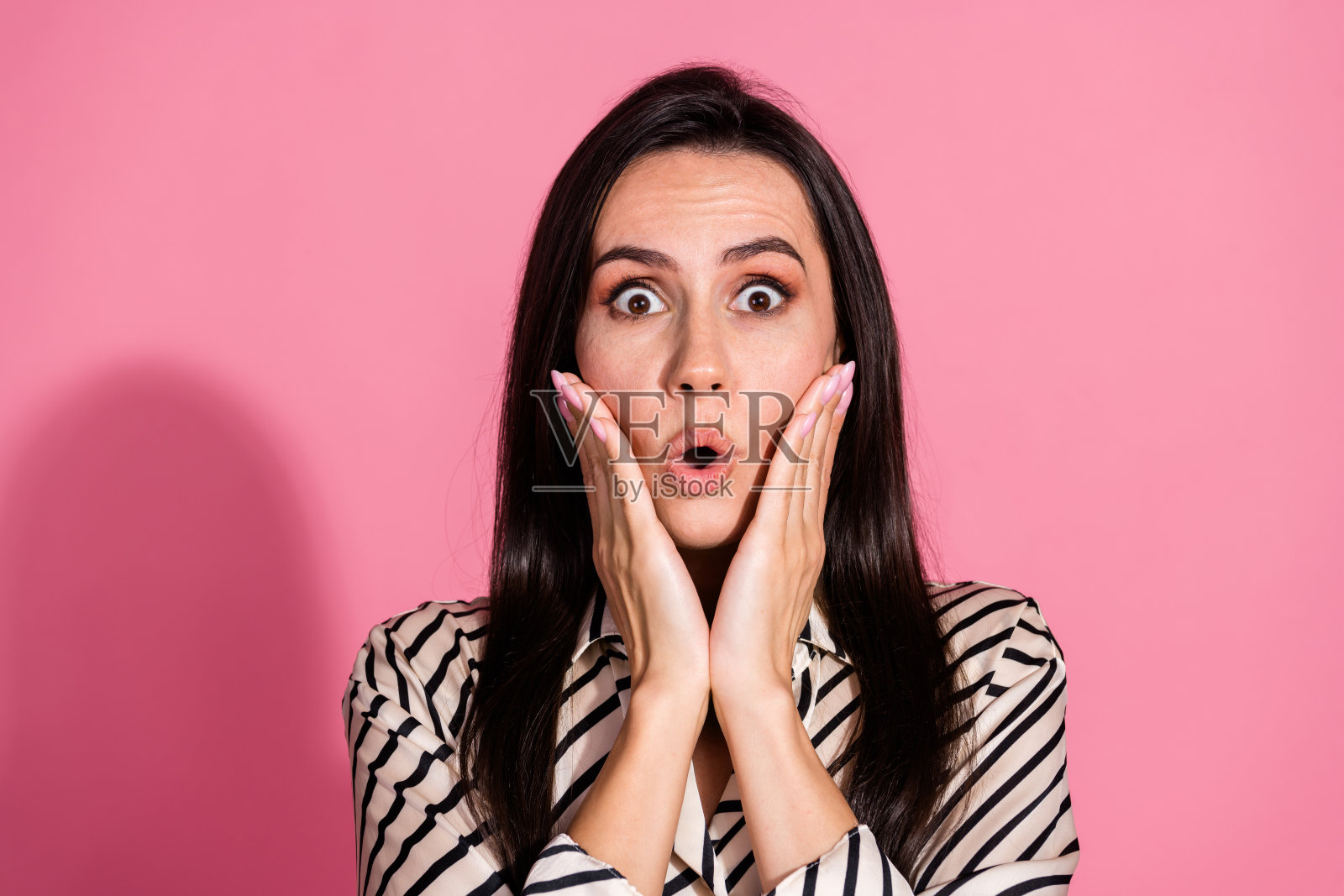 穿着条纹衬衫的年轻女性在粉色背景前露出惊讶的表情,眼睛睁得大大的。照片摄影图片