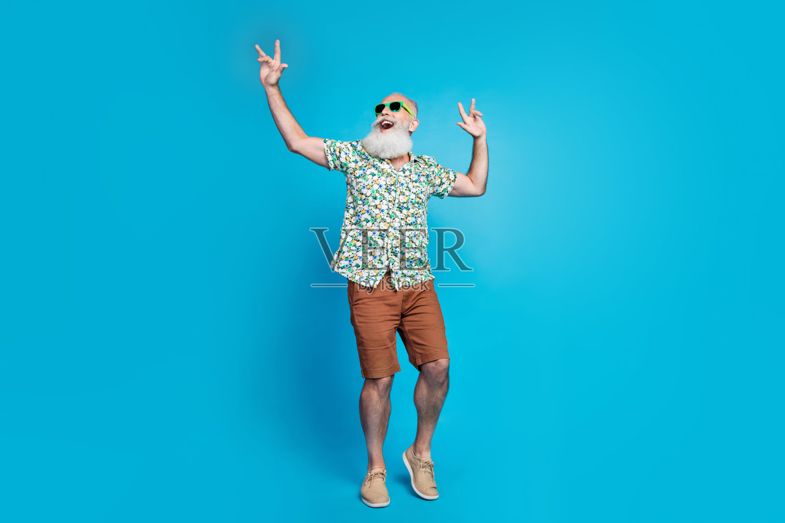 全身照,老年退休者,留着胡须和灰色小胡子,穿着热带夏季衬衫,双手摆出摇滚姿势,仰望,背景为蓝色。照片摄影图片