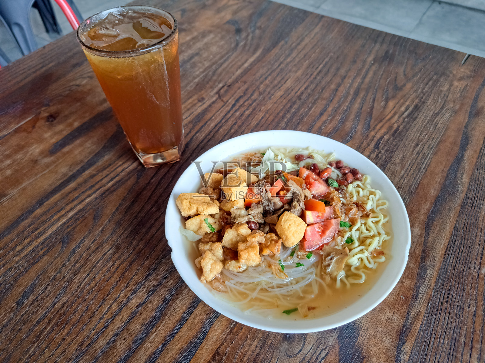 土耳其牛肉面汤,配有黄面条、米粉、牛肉、豆腐、天贝、胡萝卜、西红柿、炸洋葱、卷心菜、芹菜、炸洋葱和花生。新鲜的冰茶或玻璃杯中的清凉茶。食品和饮料菜单。照片摄影图片