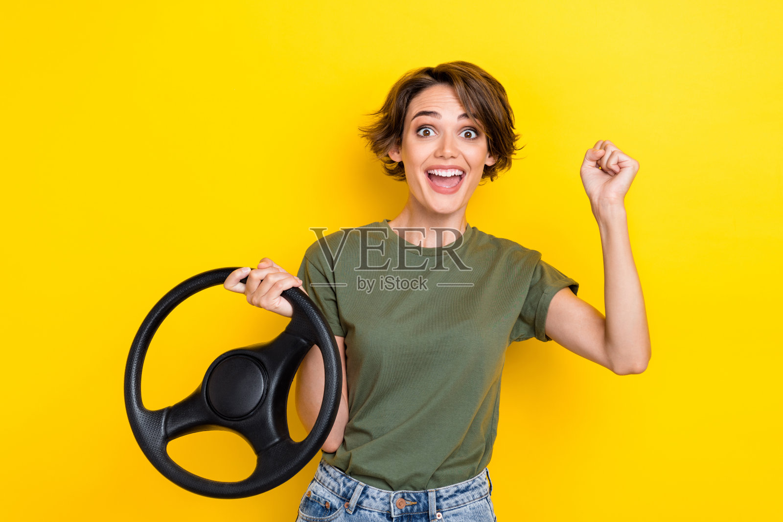 一位积极印象的女性肖像,留着波波头,穿着卡其色T恤,握着方向盘,拳头举起,背景为黄色。照片摄影图片
