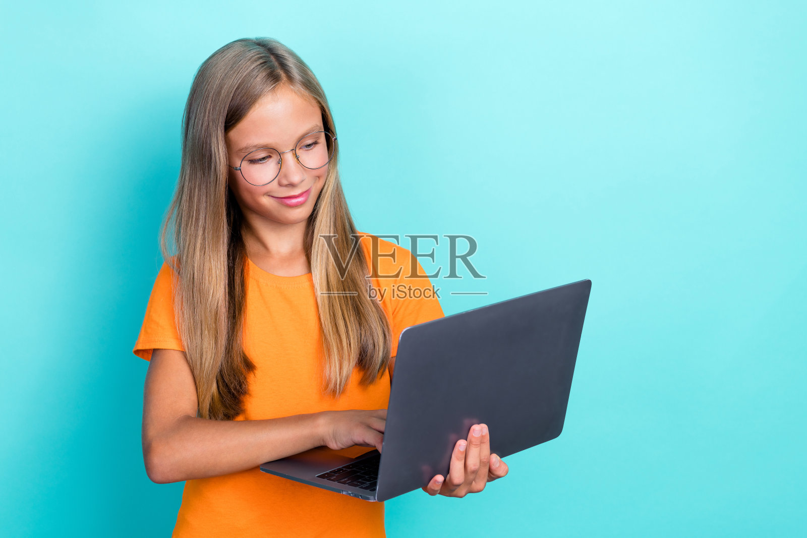 特写照片:年轻的学龄前女孩穿着橙色T恤,戴着眼镜,正在使用电脑为老师的远程教育打字,背景为青色。照片摄影图片