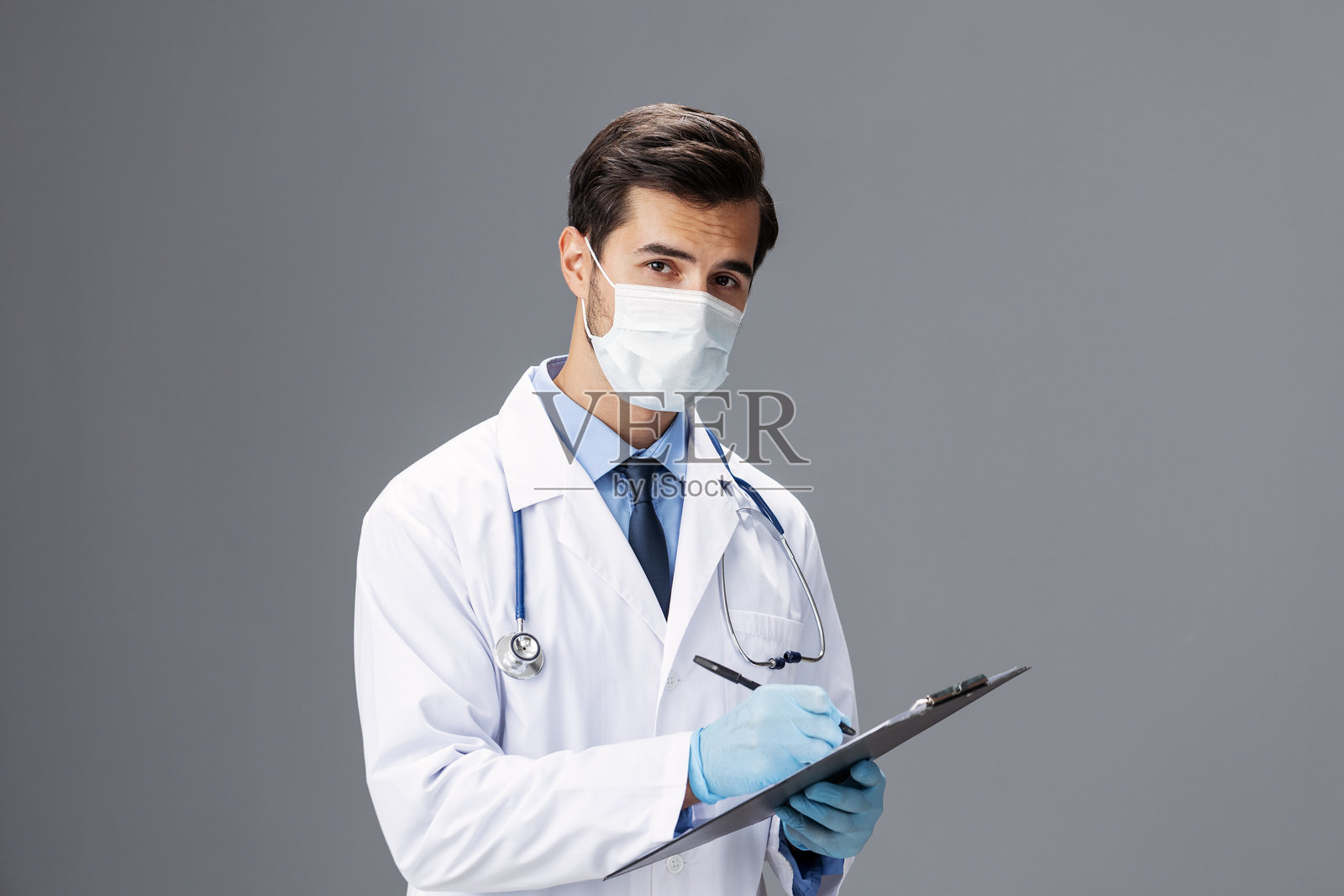 一位穿着白大褂和医用口罩的男性医生,手中拿着记事本和病历卡,正对着镜头,背景为灰色,留有空白区域,适合添加文字。照片摄影图片