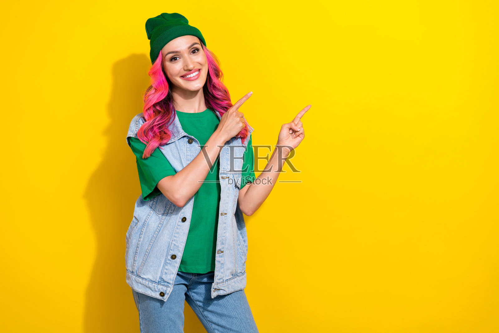 心情愉悦的美丽女孩,卷发,穿着绿色T恤,指向空旷的空间,背景为黄色。照片摄影图片