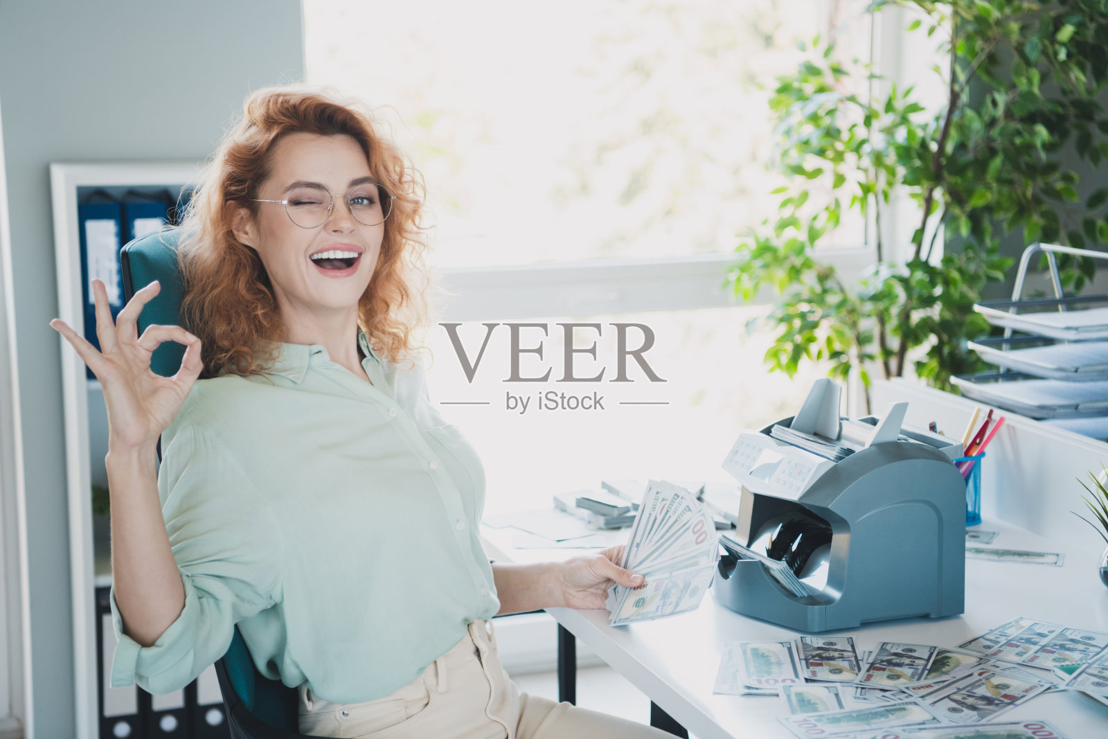 年轻的职场女性眨眼做出“OK”手势,穿着衬衫,坐在现代室内办公室里。照片摄影图片