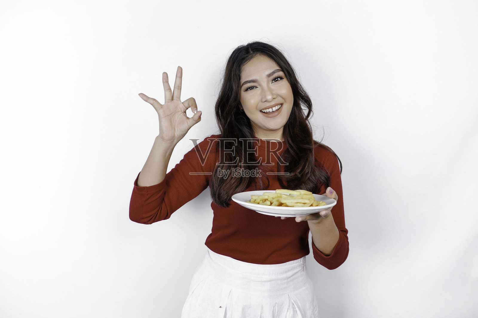 微笑的年轻亚洲女孩手持一盘薯条,做出OK手势。照片摄影图片