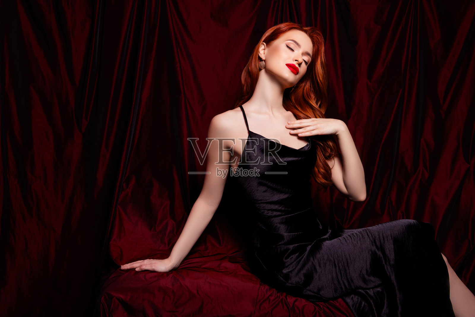 美丽迷人的女孩模特触摸柔软肌肤,穿着黑色连衣裙,背景是红色窗帘。照片摄影图片