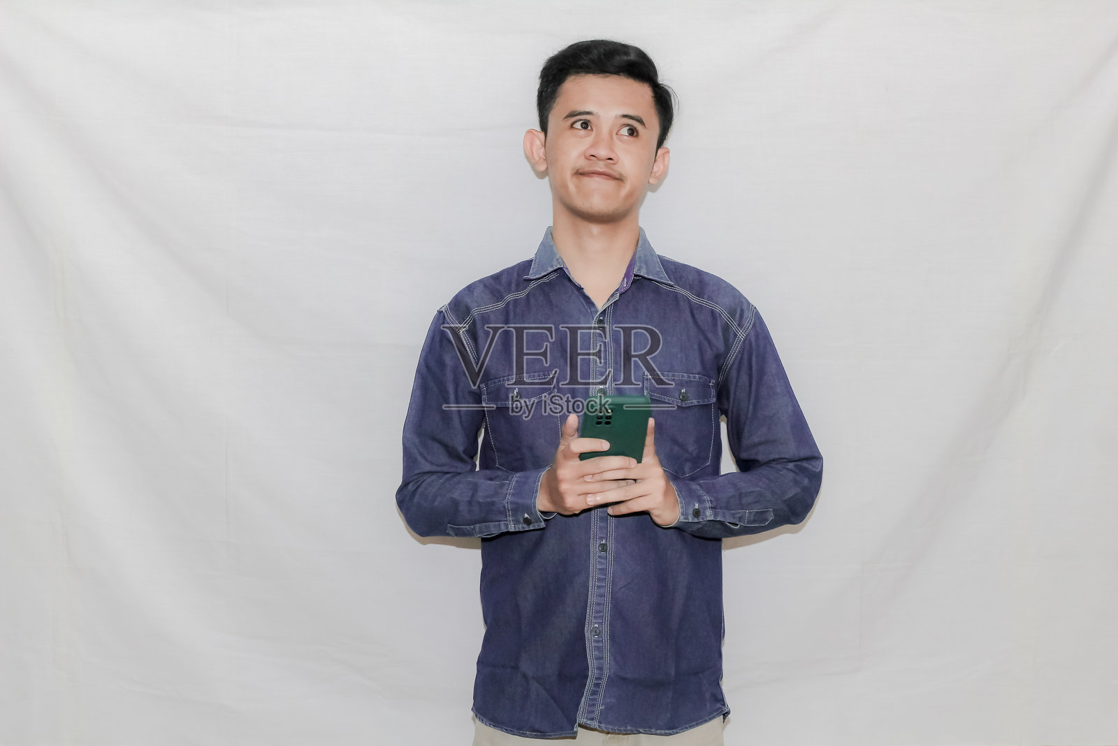 亚洲男子手持手机,表情思考某事,背景为白色。照片摄影图片