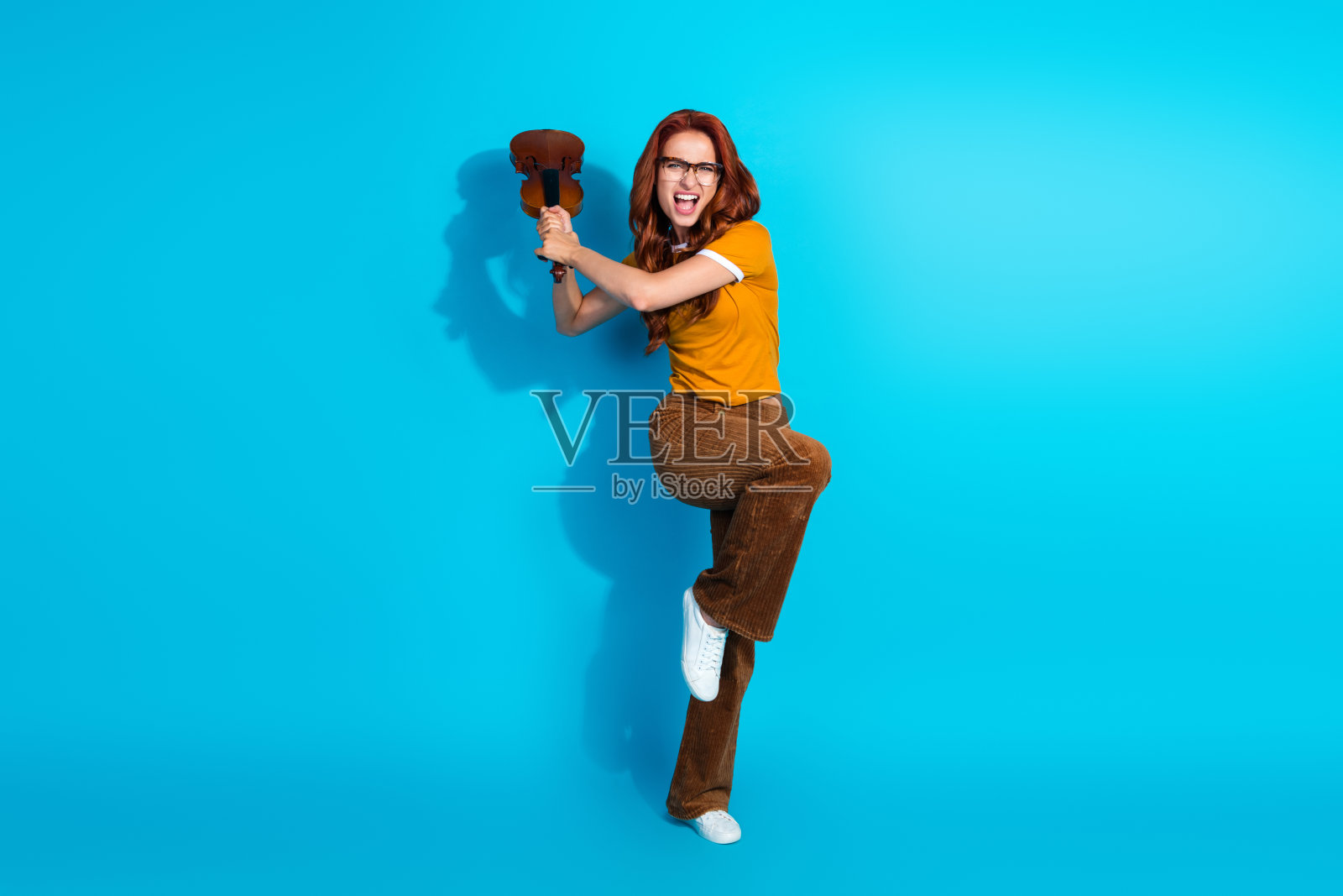 愤怒的年轻女音乐家穿着时尚服装,手持小提琴在蓝色背景下争斗的照片照片摄影图片