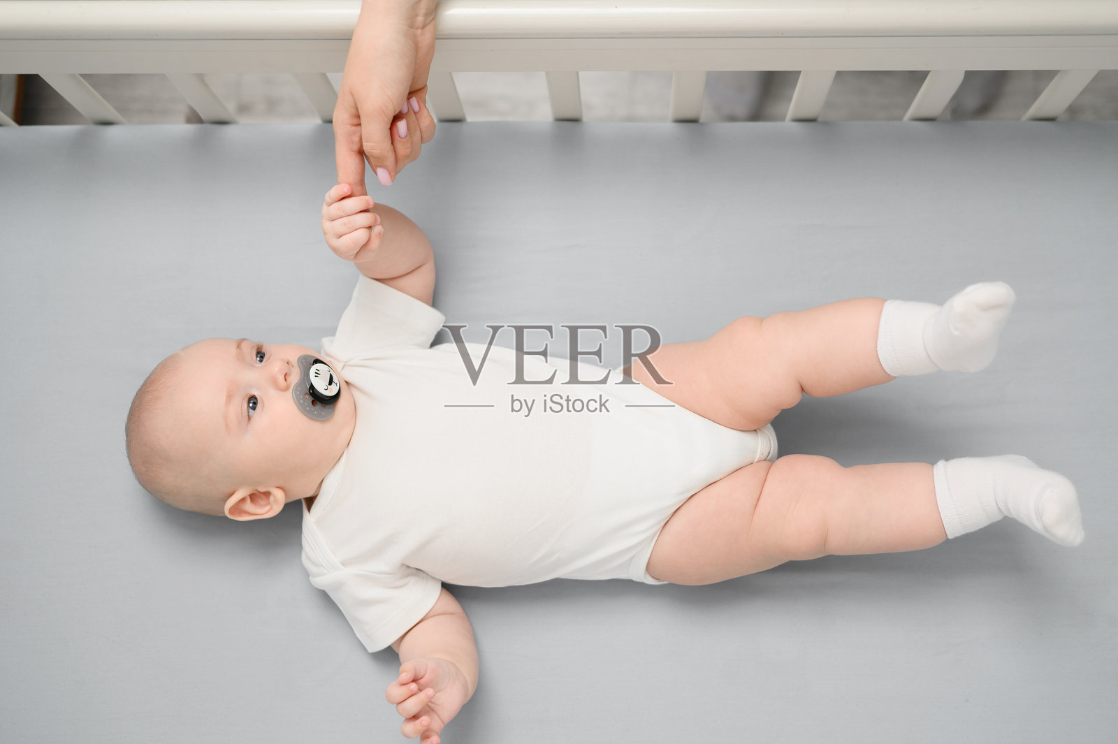 安静地躺在婴儿床里的新生儿,嘴里含着安抚奶嘴,周围是温馨的育儿环境,手握着妈妈的手,俯视角度。母亲、婴儿和产假幸福的概念。照片摄影图片