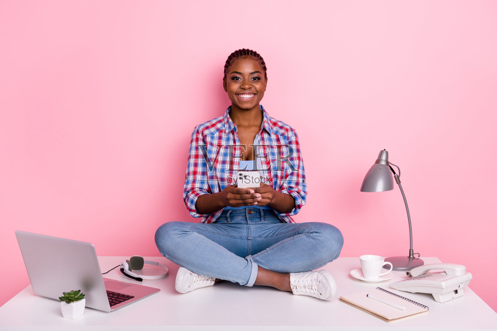 全身肖像,漂亮积极的女士坐在桌子旁,手持电话,背景为粉色。照片摄影图片