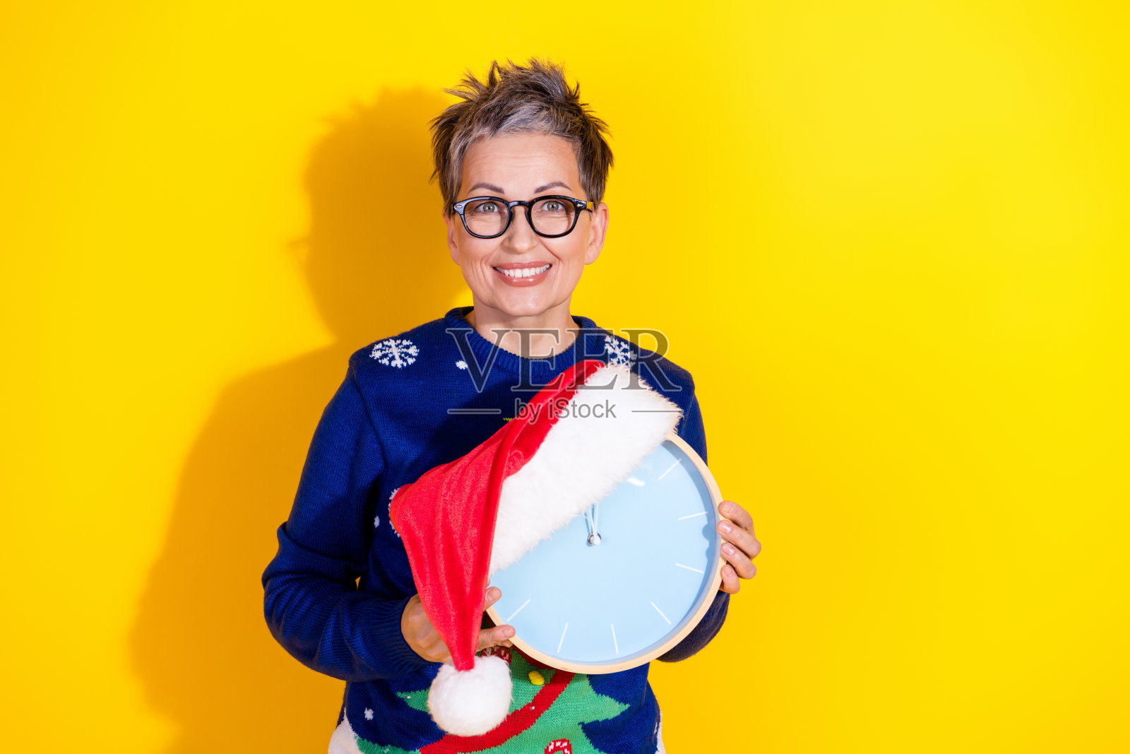 可爱的老年女士穿着蓝色时尚服装,手持时钟,等待新年夜,背景为黄色。照片摄影图片