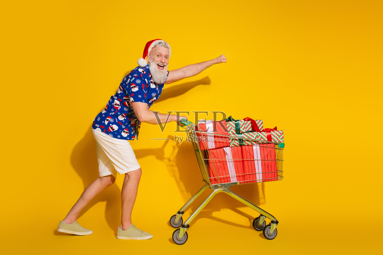 时尚圣诞老人全身肖像,推着礼品盒,穿着圣诞印花衬衫,背景为黄色。照片摄影图片