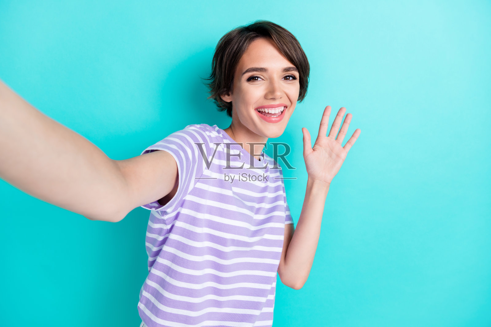 一位快乐迷人的女孩,留着波波头,穿着条纹T恤,挥舞着手掌自拍,背景为青绿色。照片摄影图片