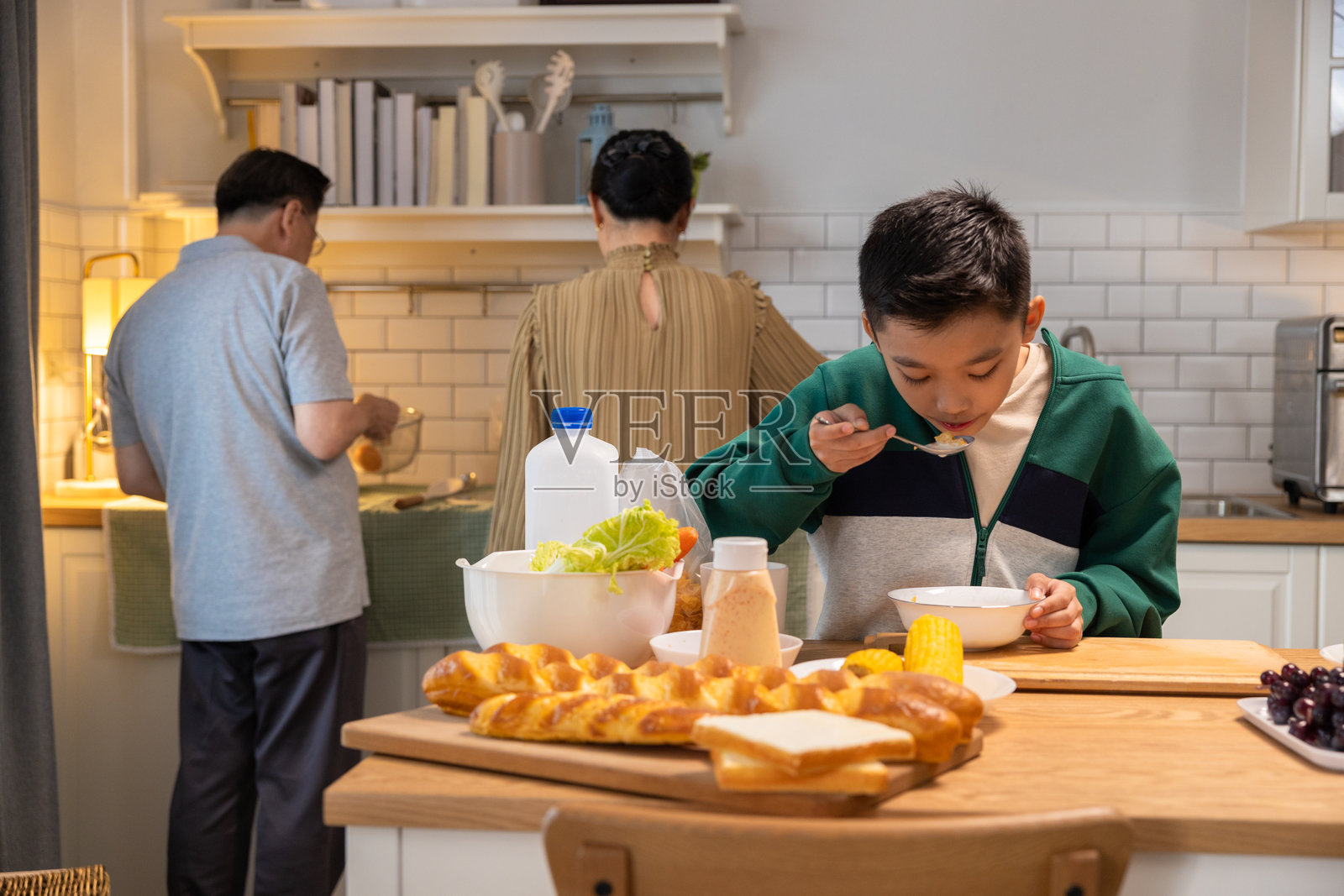 儿子在厨房吃早餐,父母正在做饭。照片摄影图片