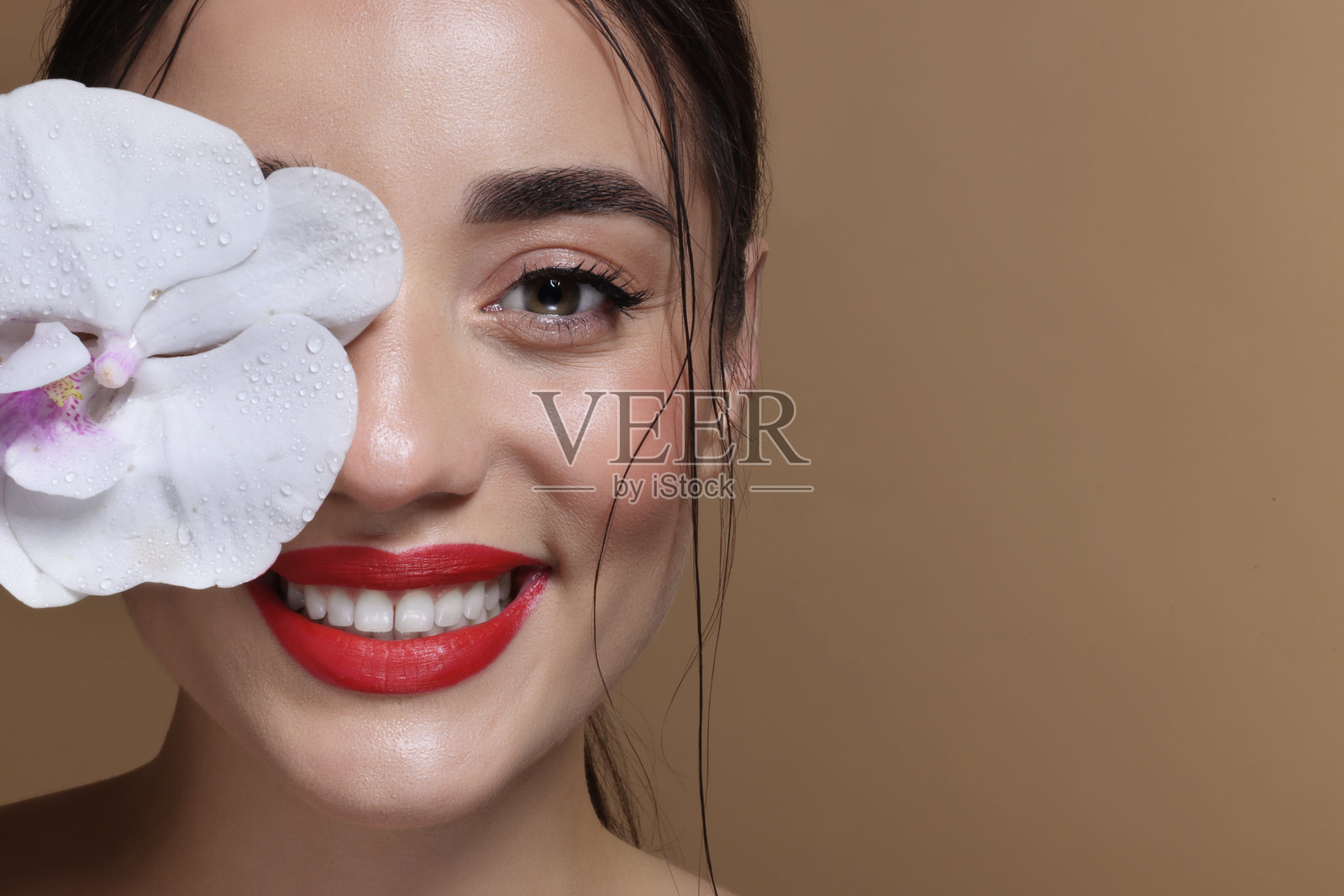 微笑女性用兰花遮住眼睛的肖像,背景为棕色,特写。留有文字空间。照片摄影图片