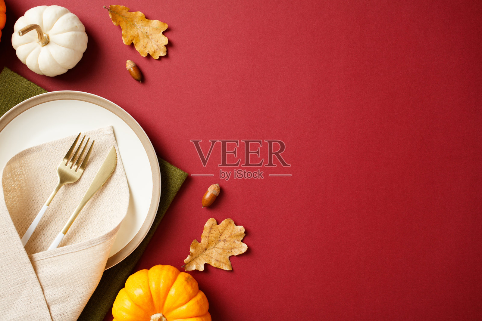 感恩节餐桌布置,白色盘子、米色餐巾、金色刀叉,秋季装饰包括南瓜、橡果和枫叶,背景为浓郁的酒红色。照片摄影图片
