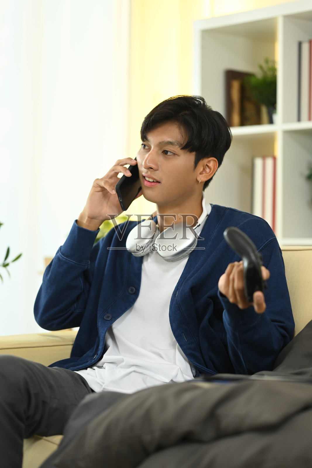 年轻男子在客厅里拿着游戏手柄并在手机上通话。照片摄影图片