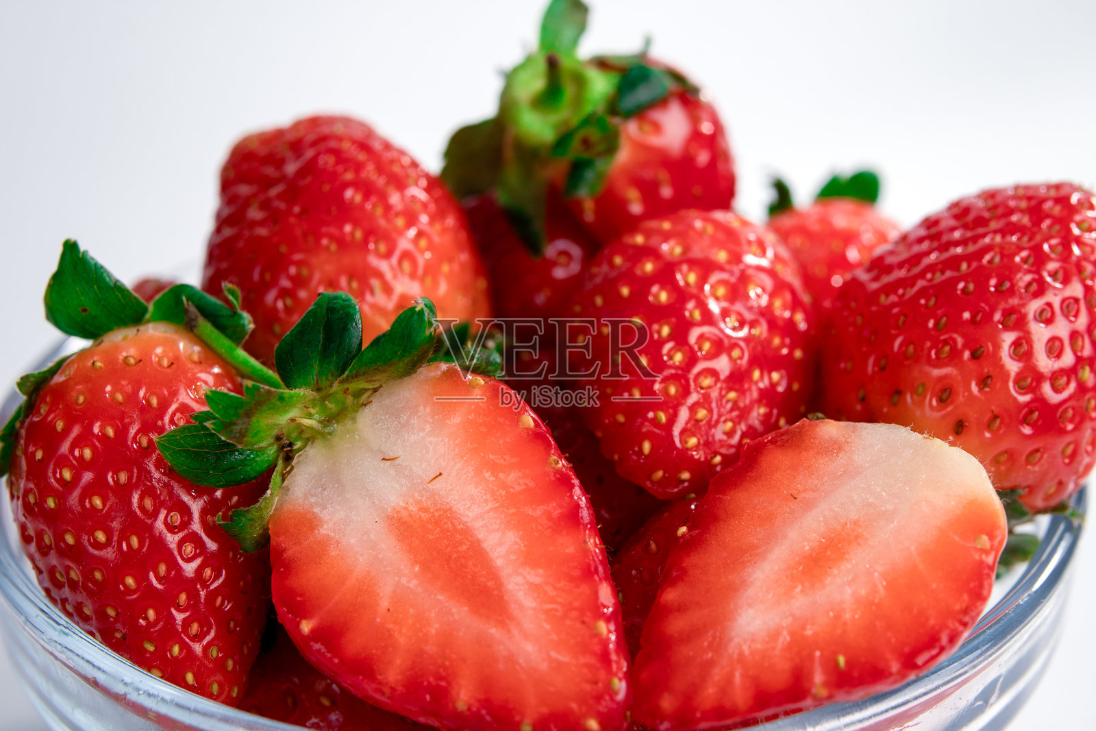 一个装满新鲜草莓的玻璃碗,里面有完整的草莓和一个切成两半的草莓,鲜艳的红色和多汁的质感在白色背景下显得格外诱人。非常适合健康、营养和食品摄影主题。照片摄影图片