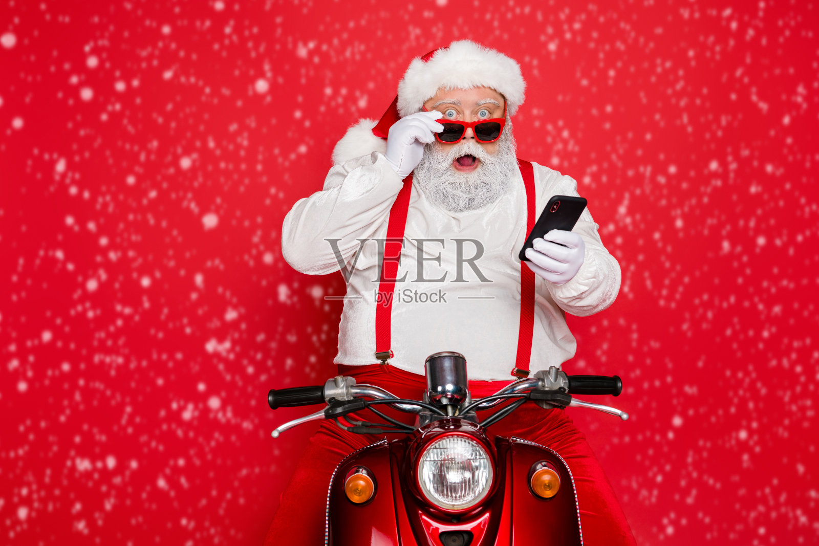 可爱的有胡子的圣诞老人,惊讶而搞笑,戴着帽子,坐在摩托车上,使用手机阅读冬季新闻,背景是明亮鲜艳的红色。照片摄影图片