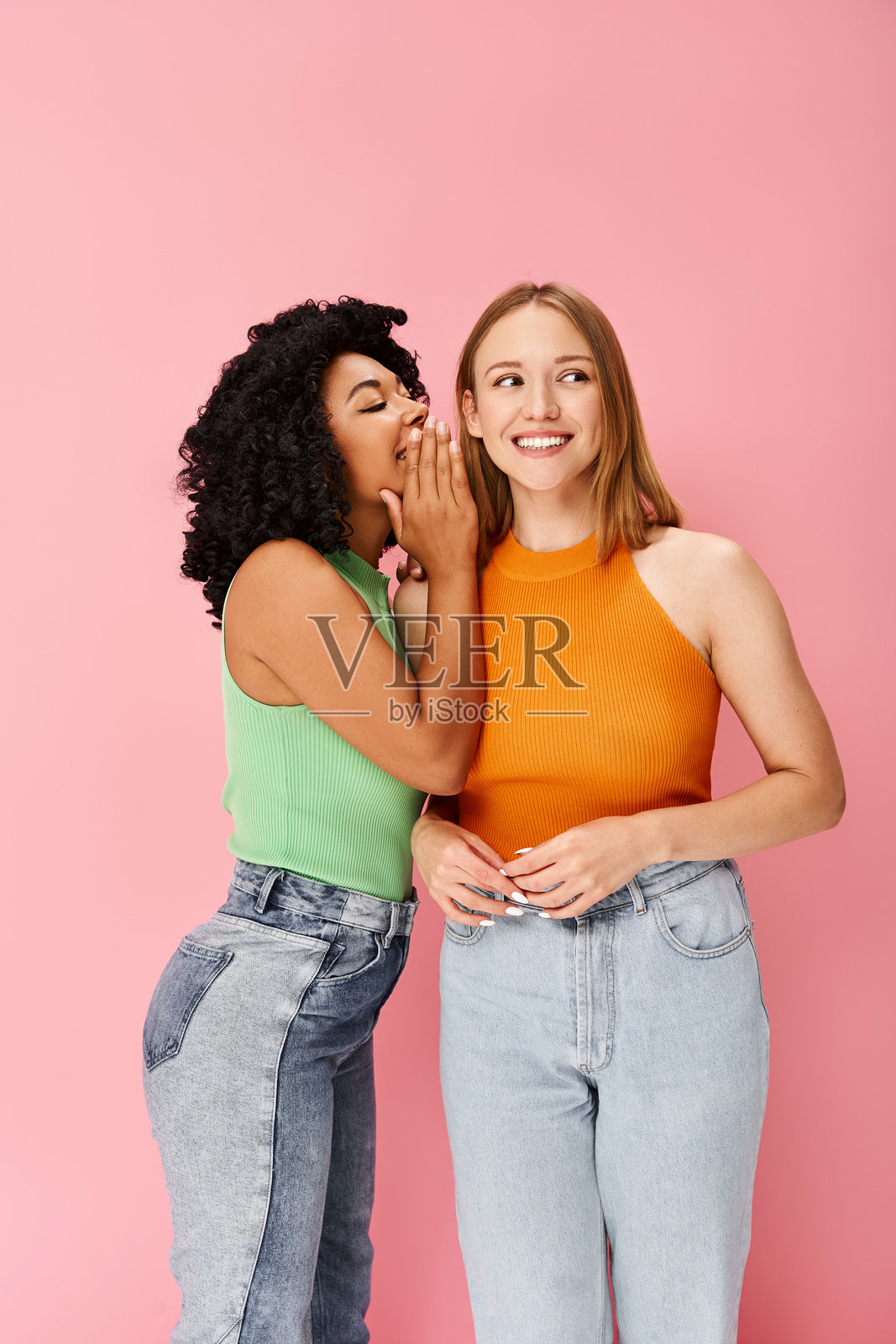 两位时尚女性在鲜艳的粉色背景下分享快乐的时刻,身穿色彩斑斓的服装。照片摄影图片