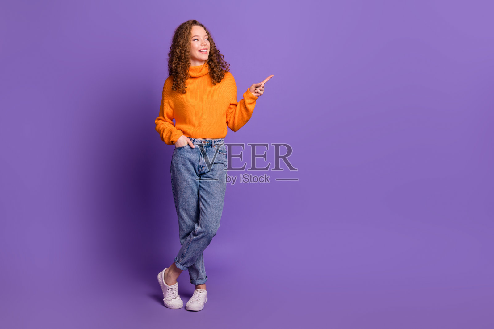 全身照,漂亮的年轻女孩指着空白处,穿着毛衣,背景为紫色。照片摄影图片