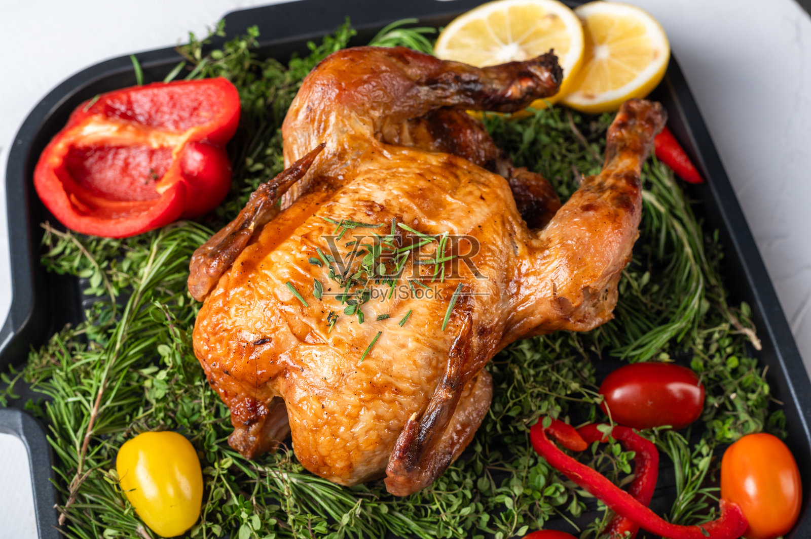 一只金黄色的烤鸡,撒上香草,放在托盘上,周围环绕着新鲜的迷迭香、百里香、柠檬片、红黄彩椒和樱桃番茄。非常适合食品博客使用。照片摄影图片