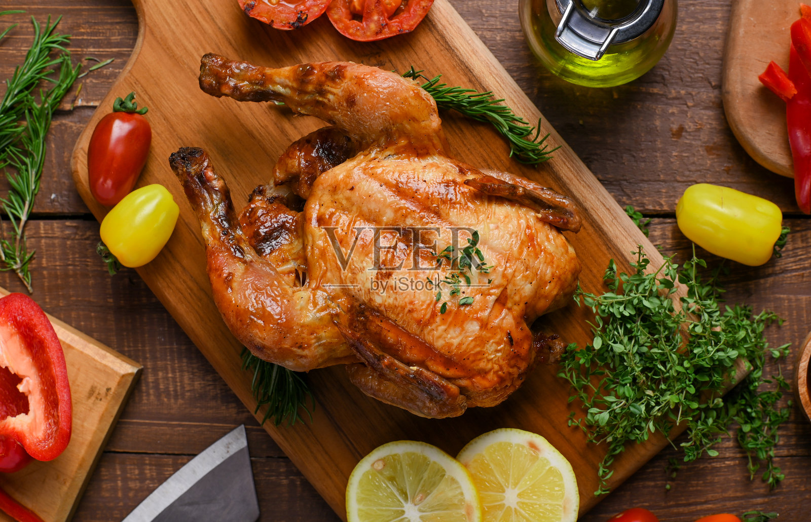 一只完美烤制的整只鸡,配以香草装饰,放在木板上,旁边有切片柠檬、红黄彩椒、烤番茄、新鲜迷迭香和橄榄油。照片摄影图片