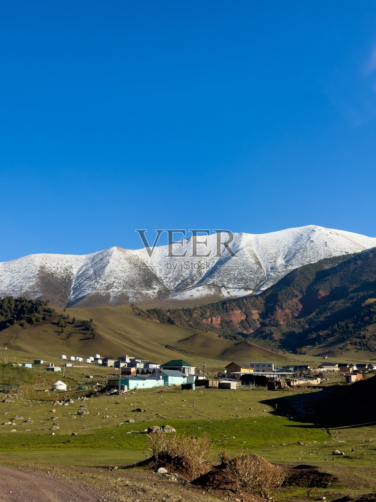 吉尔吉斯斯坦一座小村庄,坐落在雪山脚下,四周环绕着绿野和巨石,蓝天清澈。照片摄影图片