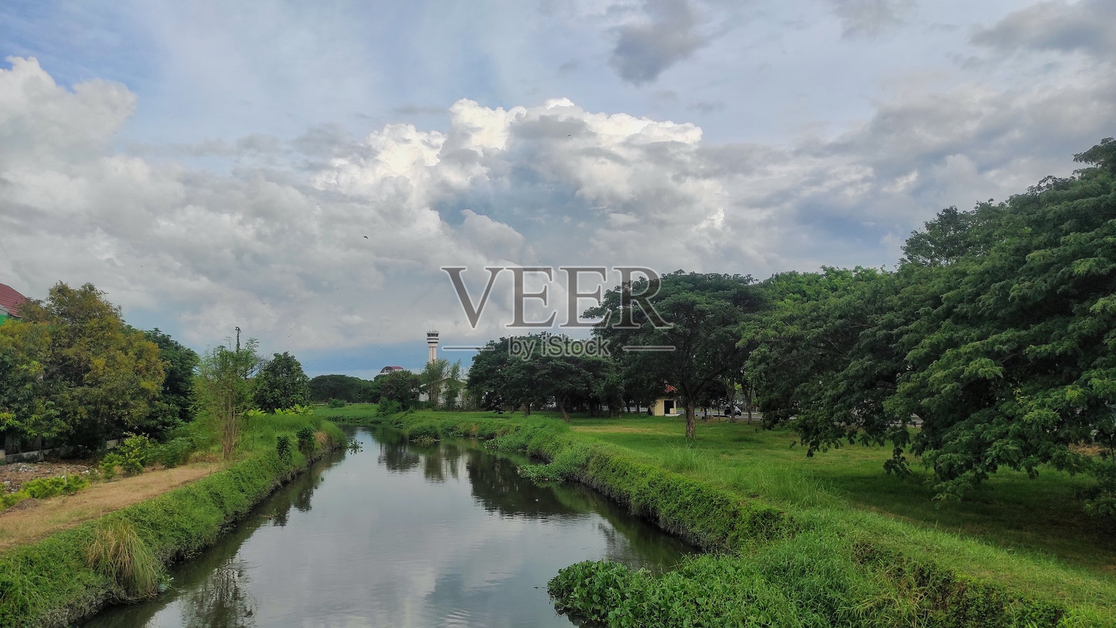 宁静的河边景色,郁郁葱葱的绿树在阴云密布的天空下。平静的河水映照着周围的树木,远处矗立着一座高塔,为这幅宁静的风景增添了深度。照片摄影图片