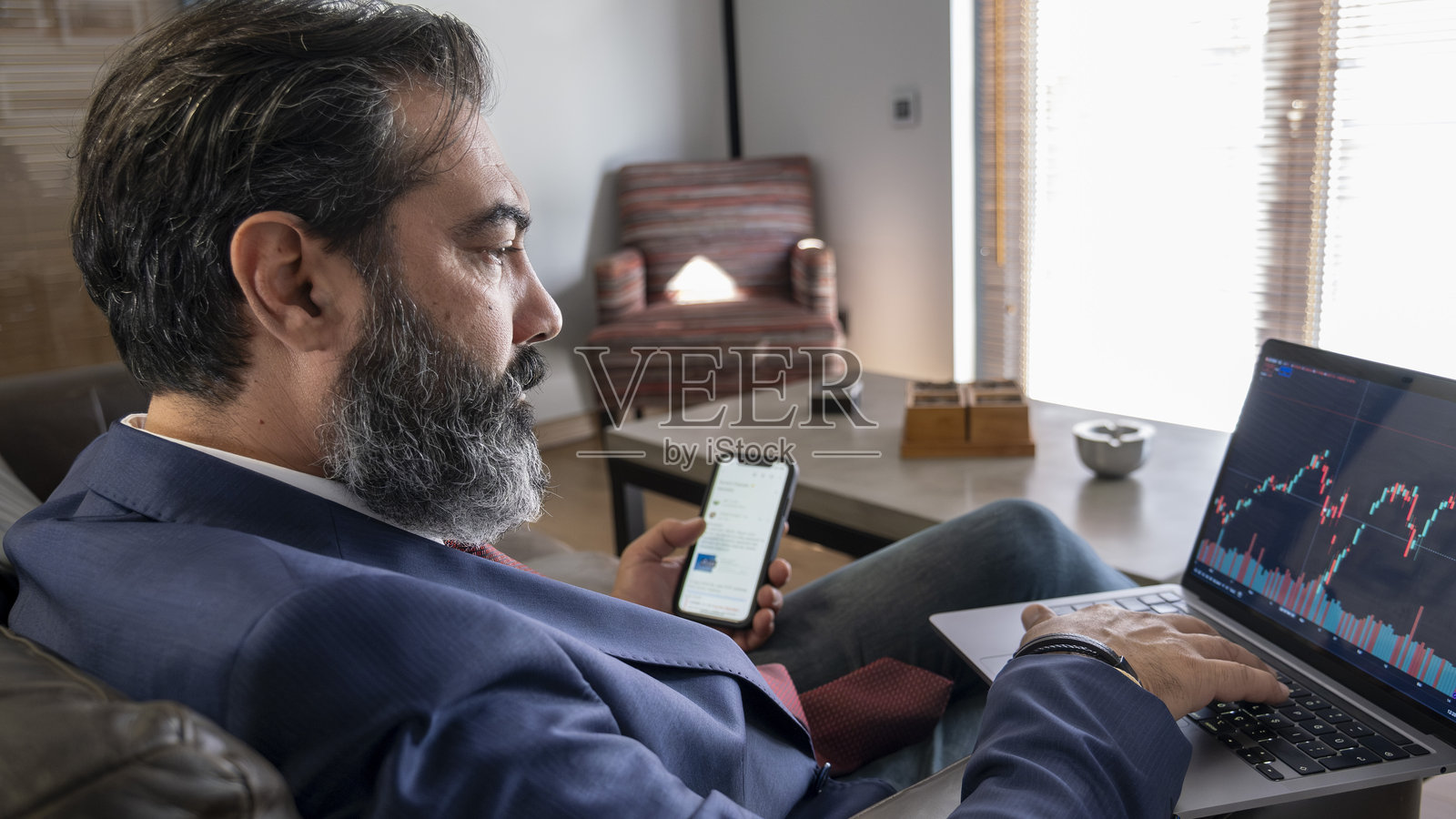 一位中年黑发商人在办公室的皮沙发上坐着,监控图形并在笔记本电脑上工作。照片摄影图片