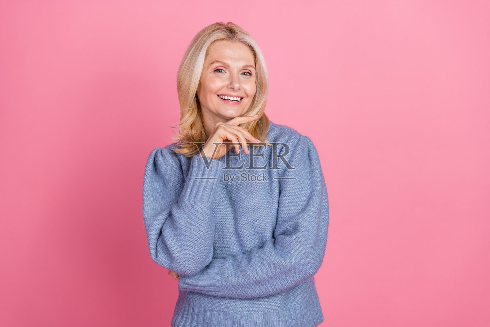 迷人的老年女性肖像,思考时轻触下巴,穿着时尚的蓝色针织服装,背景为粉色。照片摄影图片