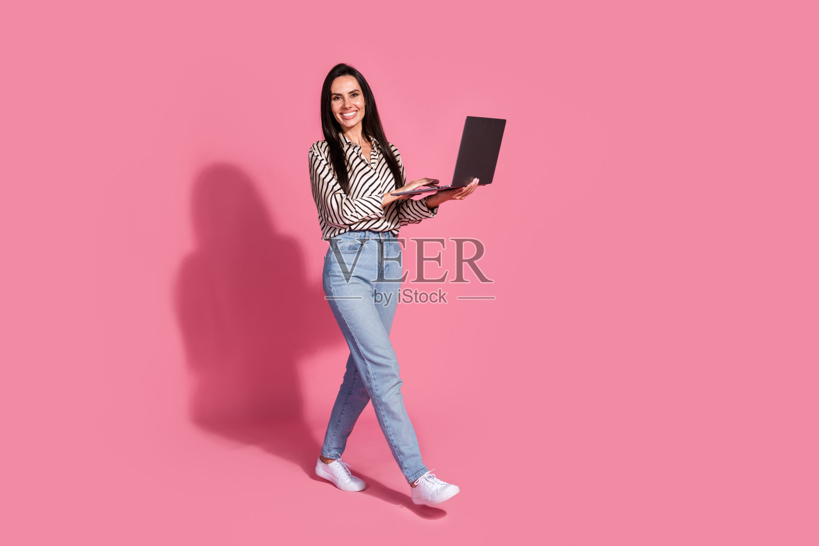 自信的年轻女性,棕色头发,穿着休闲条纹衬衫和牛仔裤,坐在粉色背景前使用笔记本电脑。照片摄影图片