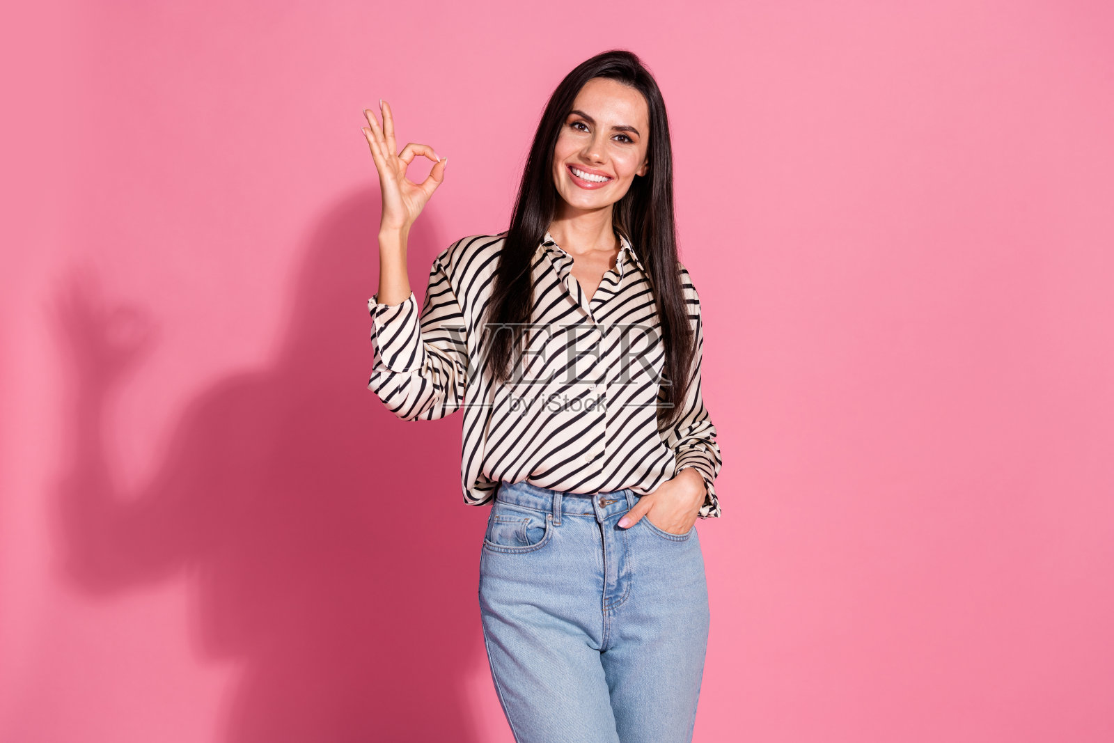 自信的年轻女性,棕色头发,穿着休闲条纹衬衫和牛仔裤,站在粉色背景前。照片摄影图片