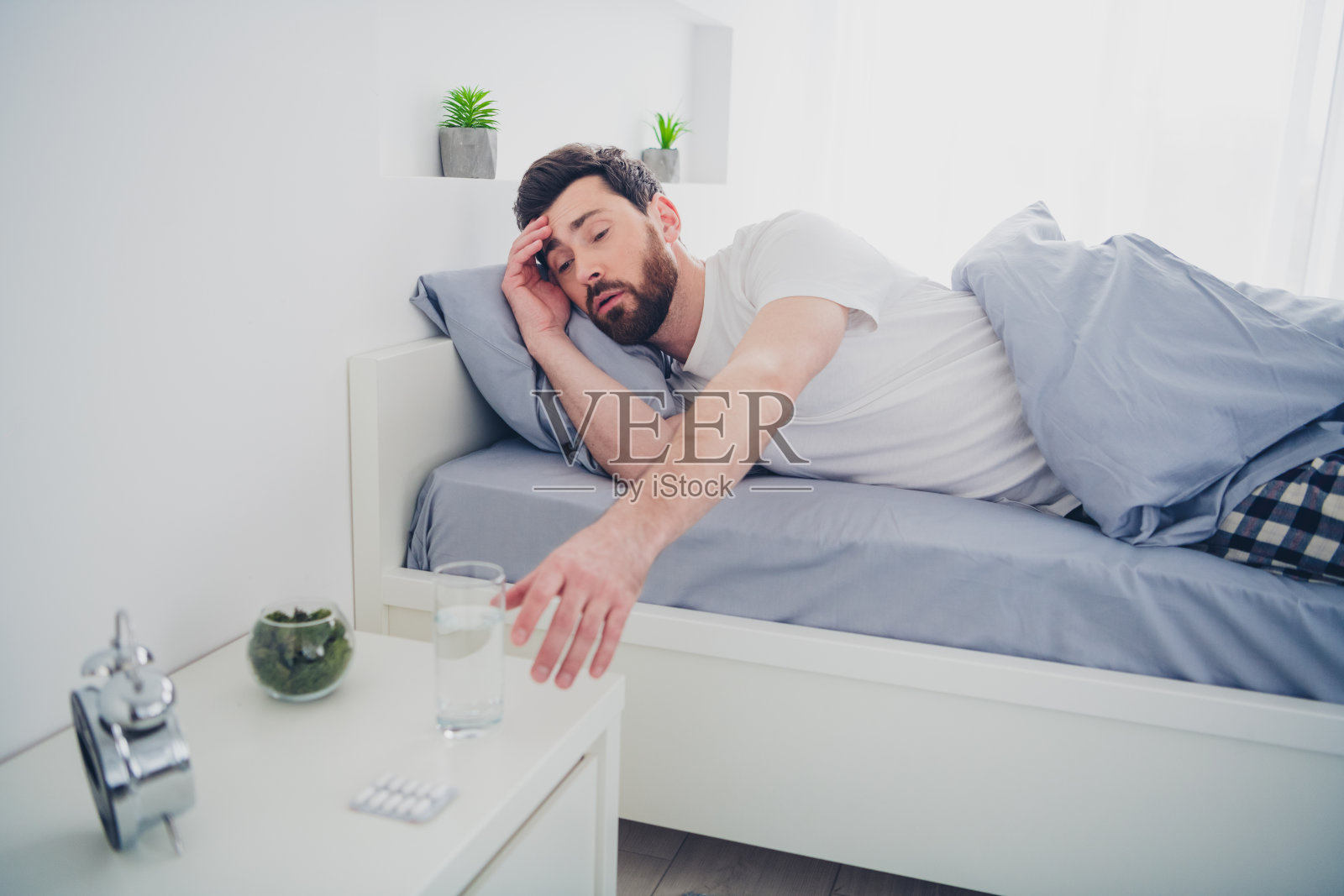 一位穿着睡衣的搞笑疲惫年轻男子躺在床上,想要喝水,室内公寓房间。照片摄影图片