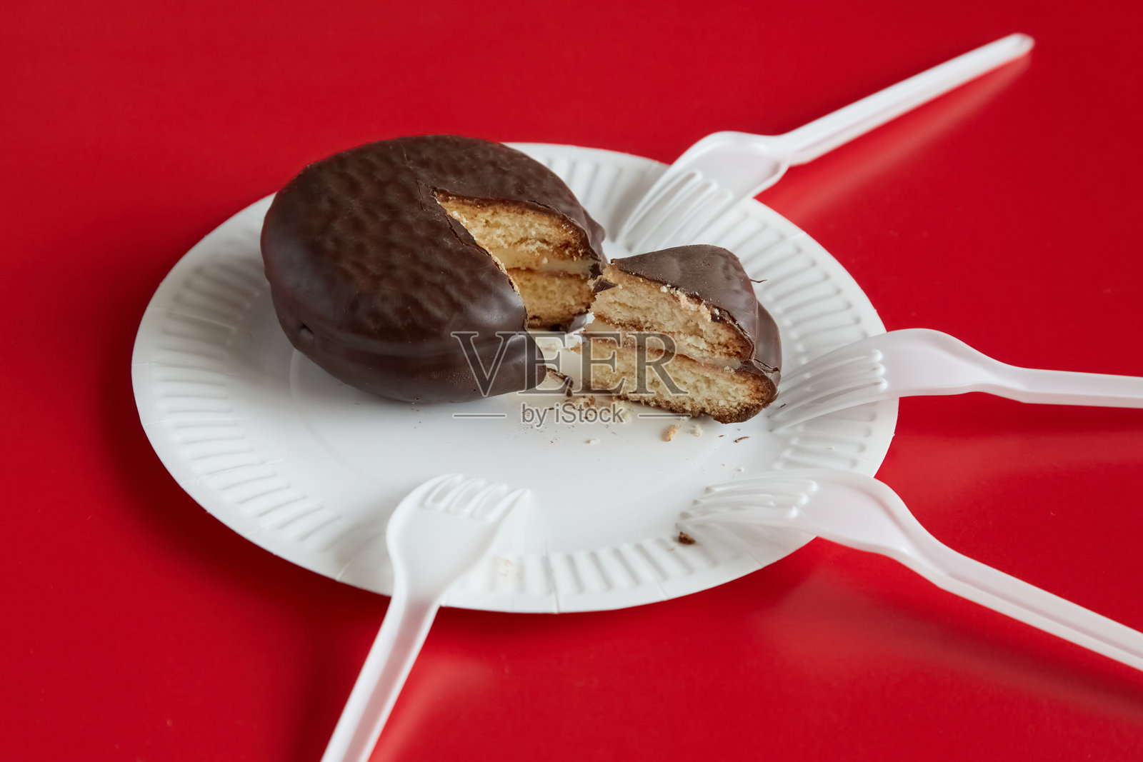 一块切好的巧克力蛋糕放在白色一次性盘子上,背景是红色,周围有四把叉子,象征着蛋糕的分配、资源有限、稀缺和高需求。照片摄影图片