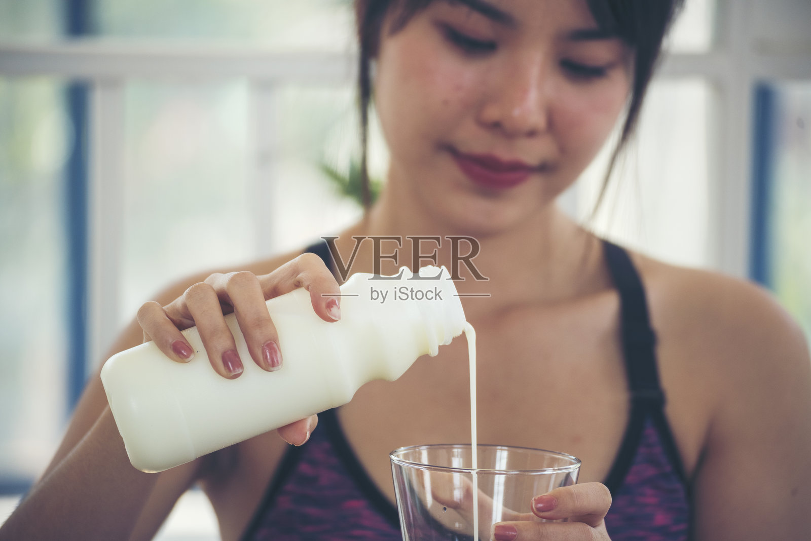 亚洲女性微笑大笑,面向镜头,健康护理家庭健身生活方式。美丽的女性饮用新鲜牛奶,低脂、无乳糖的乳制品。女性手持白色牛奶杯,从瓶子中倒出牛奶。照片摄影图片