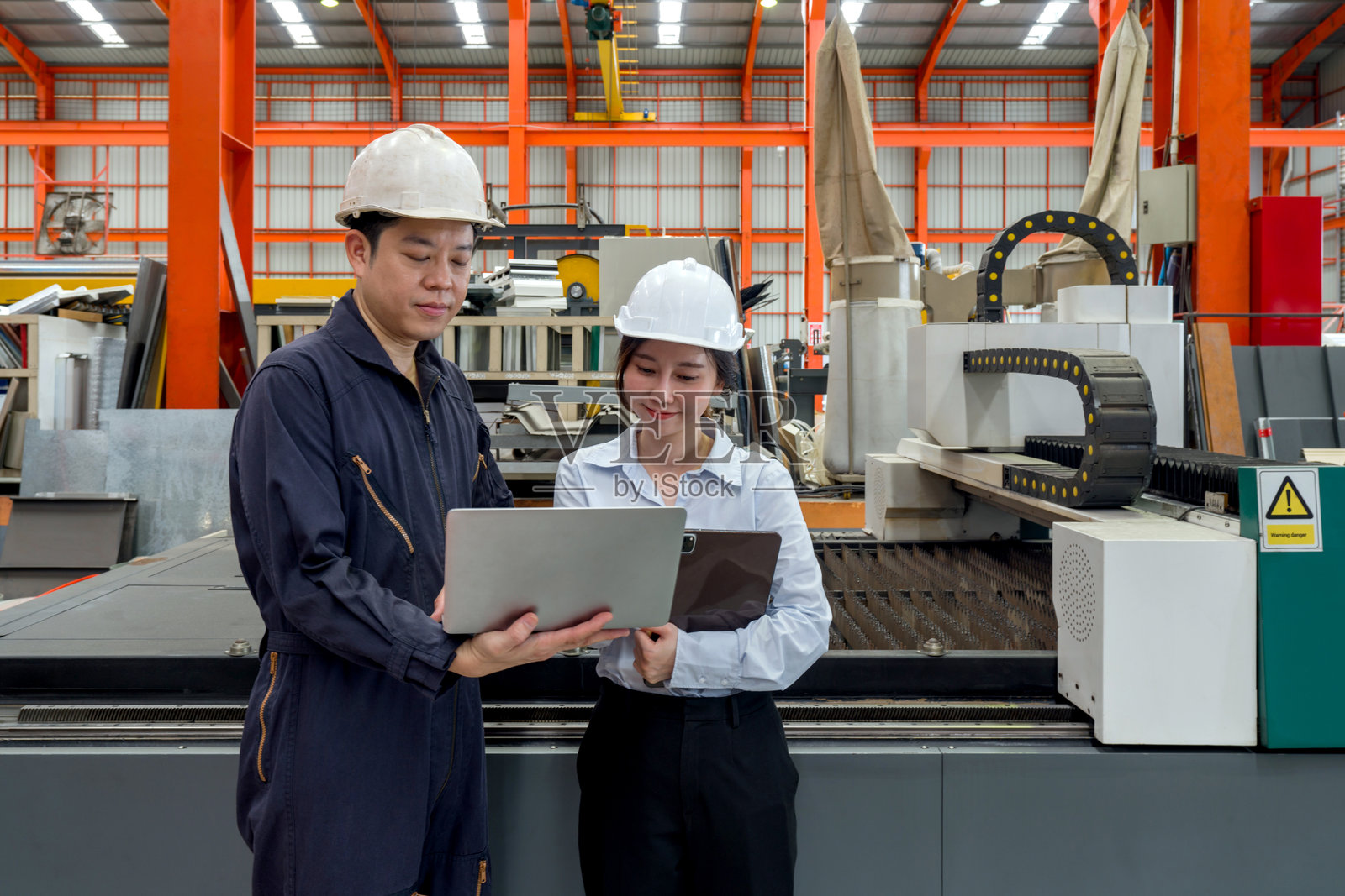 两名工人在工厂中查看笔记本电脑和平板电脑上的信息。他们戴着安全头盔,站在激光切割机前,进行高精度光纤激光切割的制造工作。照片摄影图片